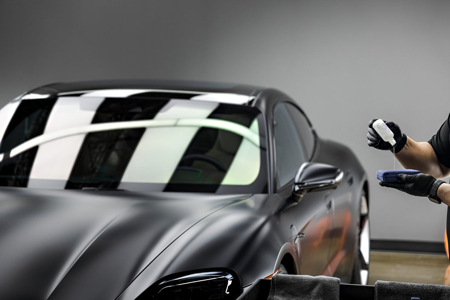 A technician in Sydney, Chipping Norton, wearing black gloves, carefully applies a ceramic coating to the matte black exterior of a Porsche Taycan. This image showcases the meticulous process of ceramic coating application, a premium service available near you. The technician is seen dispensing the ceramic coating onto an applicator pad, preparing to create a durable and protective layer on the car's paint. Consider professional ceramic coating near me to enhance and safeguard your vehicle's finish.